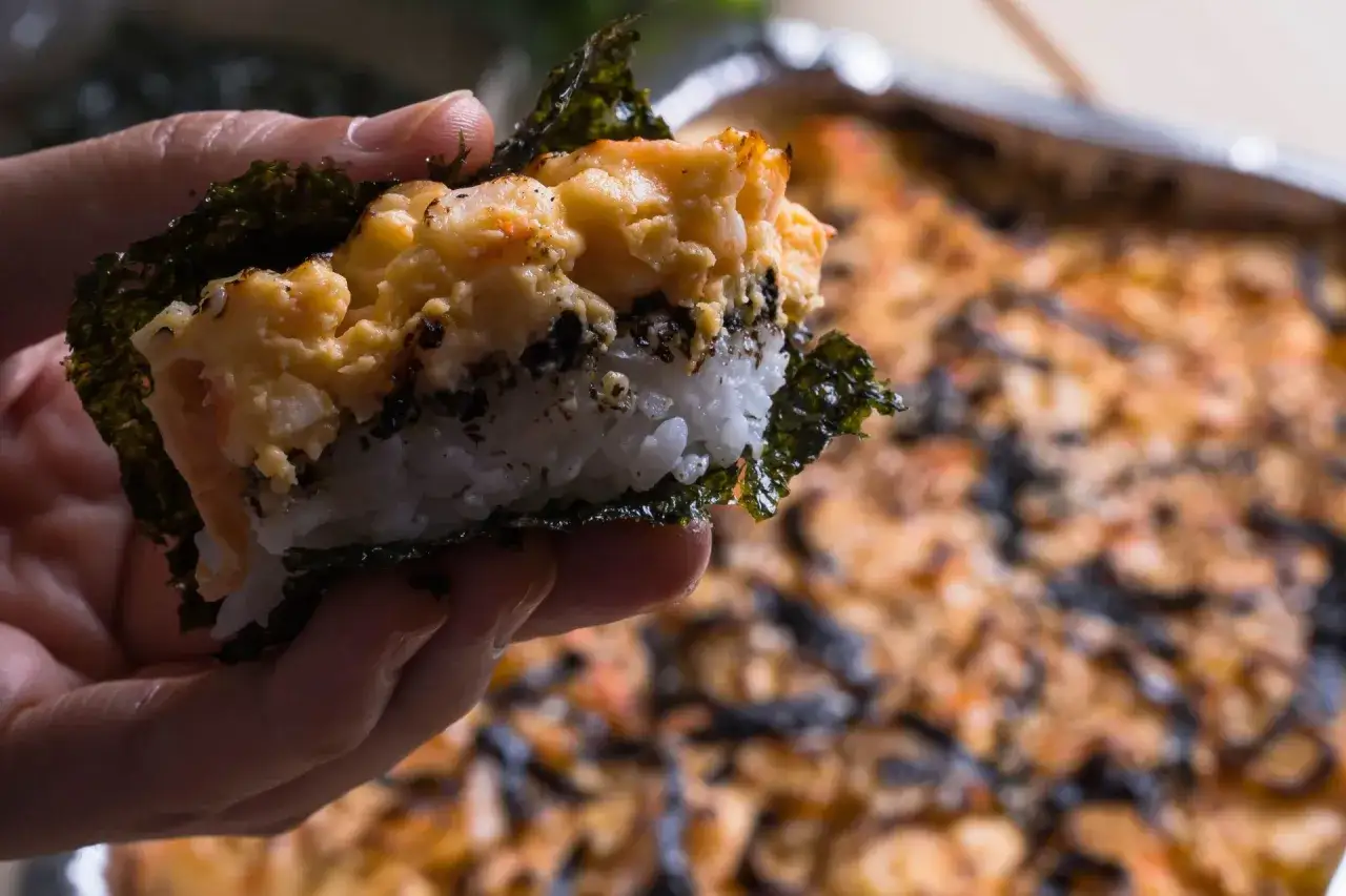 Dłoń trzyma kawałek sushi bake z ryżem, nori i zapiekanym farszem.