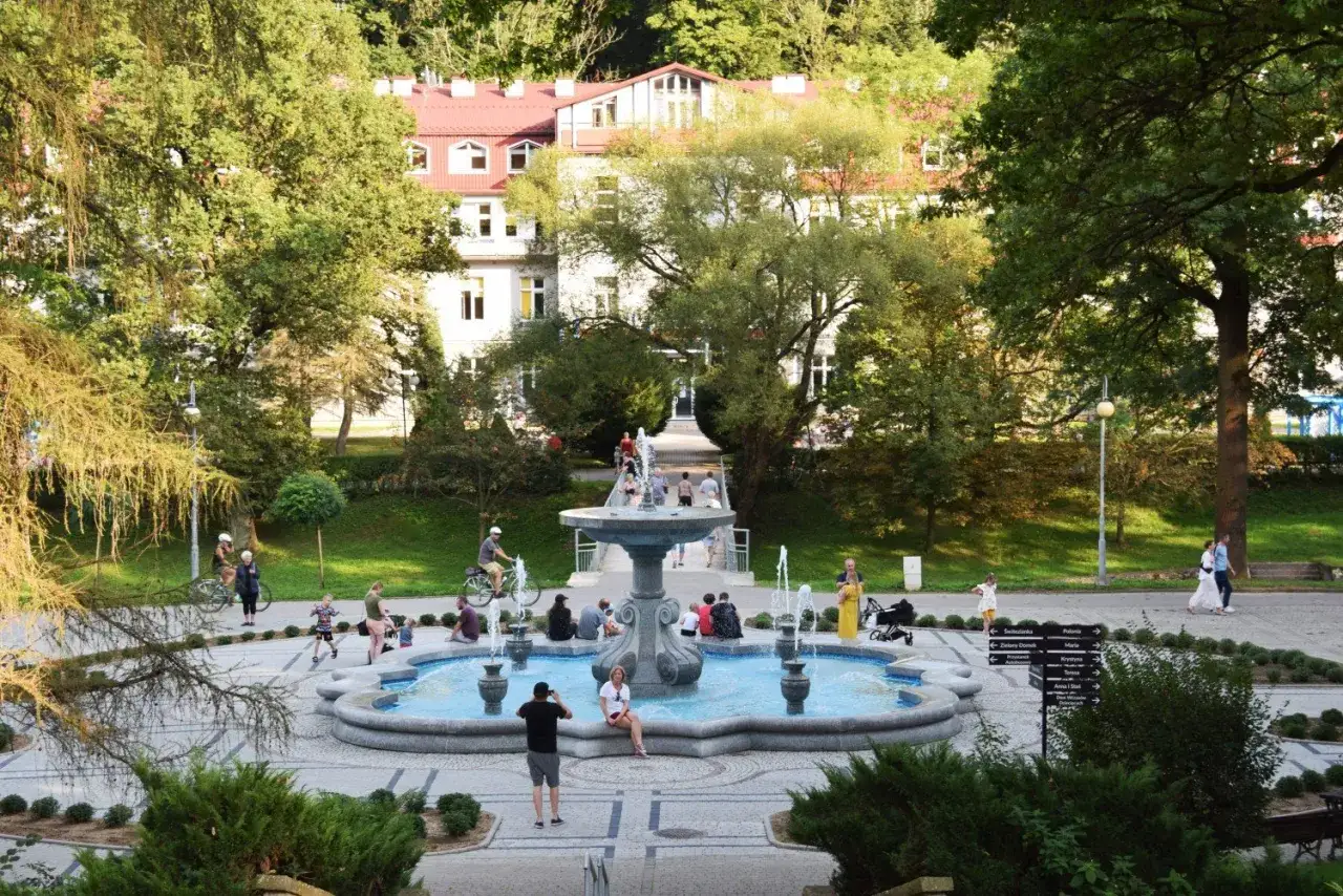 Rymanów Zdrój atrakcje: ludzie odpoczywają przy fontannie w parku zdrojowym, w tle budynek uzdrowiskowy.