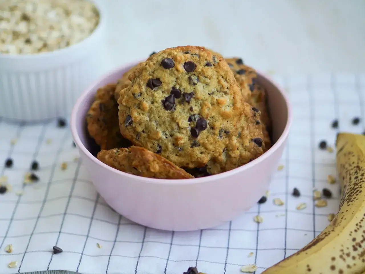 Jak zrobić ciastka dietetyczne? Pyszne ciasteczka owsiane z kawałkami czekolady w różowej miseczce, obok dojrzały banan.