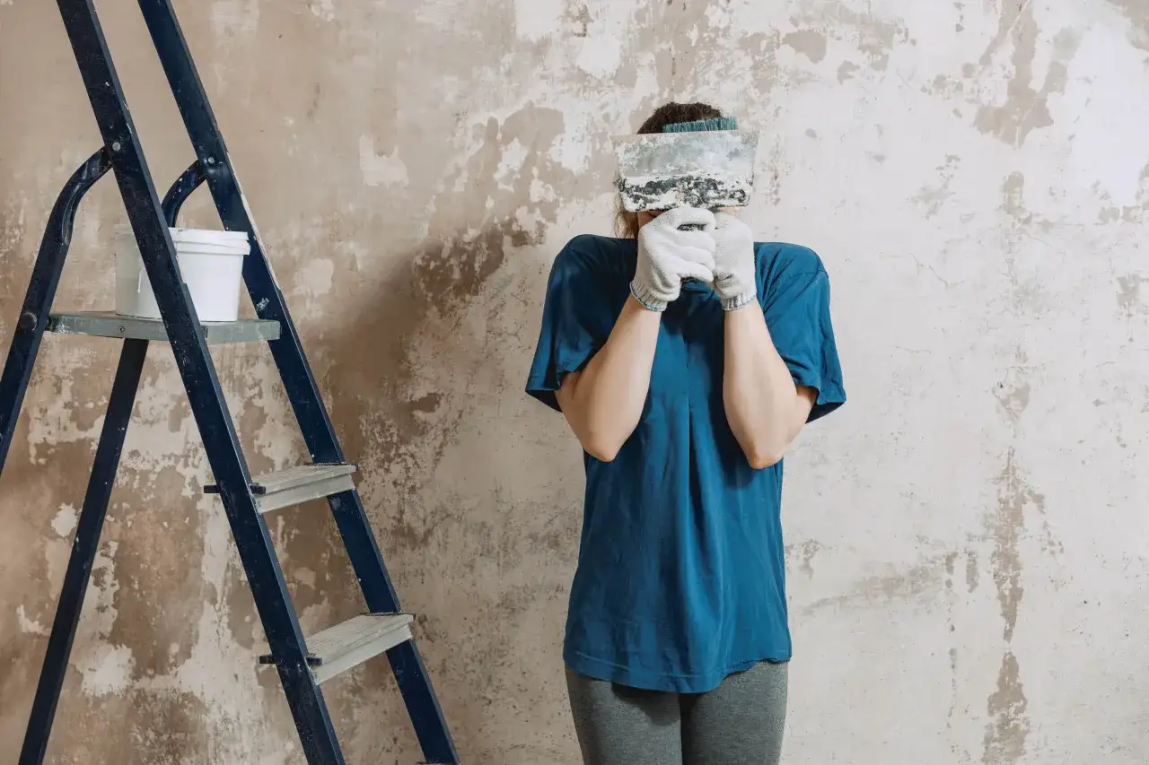 Osoba w rękawiczkach trzyma szpachelkę przed twarzą, obok drabiny z wiadrem.