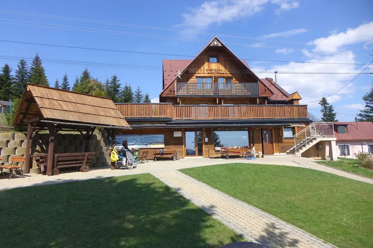 Drewniana bacówka na obidzy z balkonem i tarasem, otoczona zielenią i ścieżkami. Obok wózek inwalidzki z żółtą torbą.