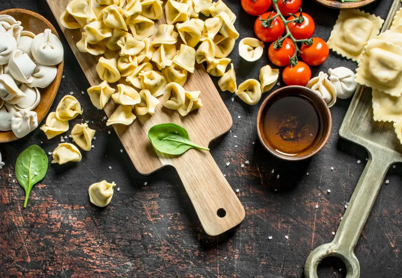 Surowe ravioli i tortellini na desce, obok pomidorków i miseczki z sosem.