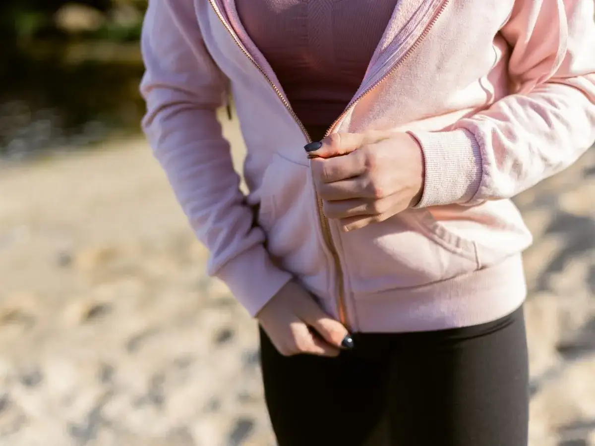 Dłoń zapina różową bluzę na zamek. Widać złote zatrzaski przy bluzie i czarne legginsy.