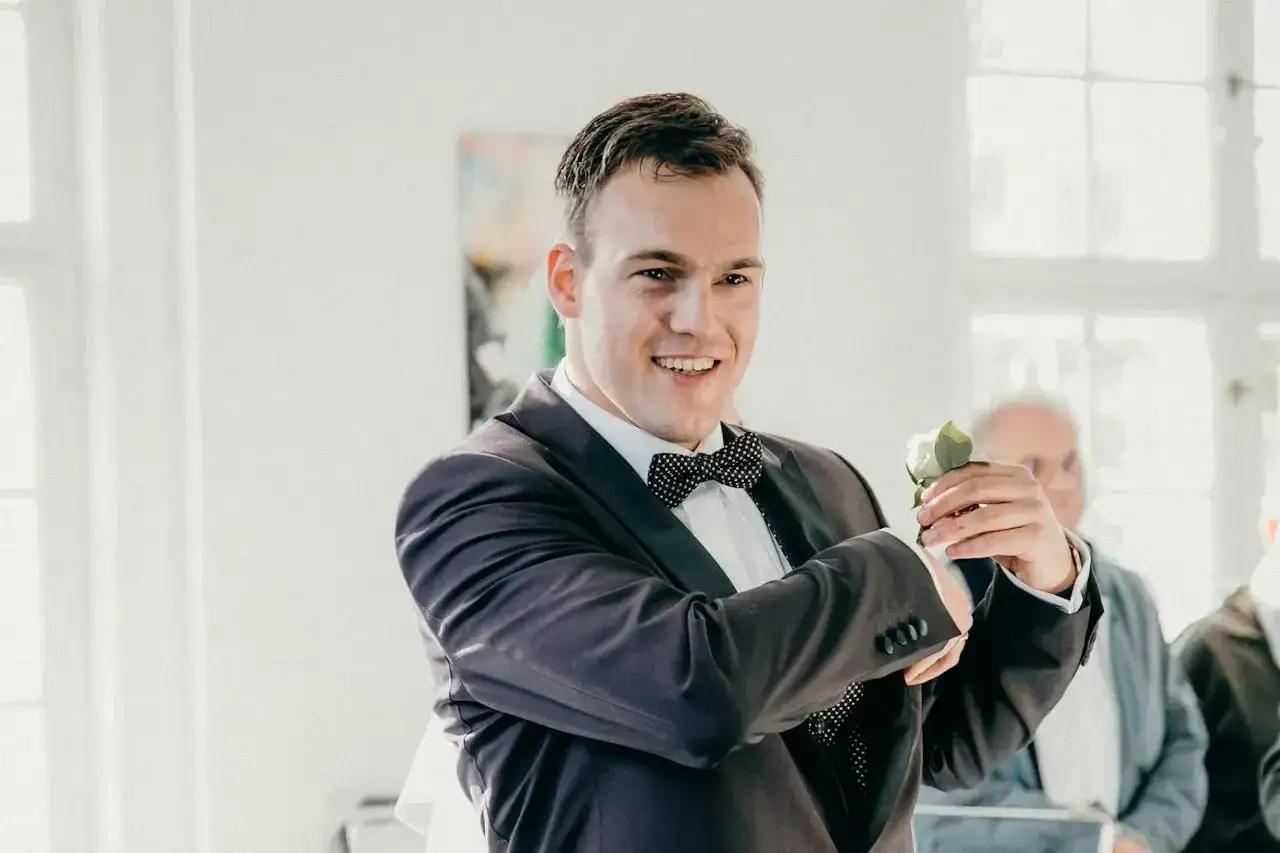 Uśmiechnięty pan młody w garniturze z muszką, trzymający różę. Zastanawia się, ile dać bratu na ślub.