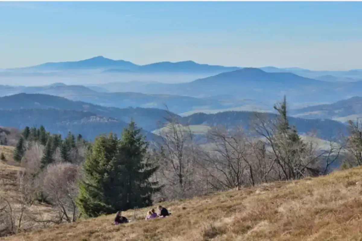 Widok na Beskid Wyspowy z kilkoma osobami odpoczywającymi na zboczu. W oddali widać kolejne pasma górskie w lekkiej mgle.