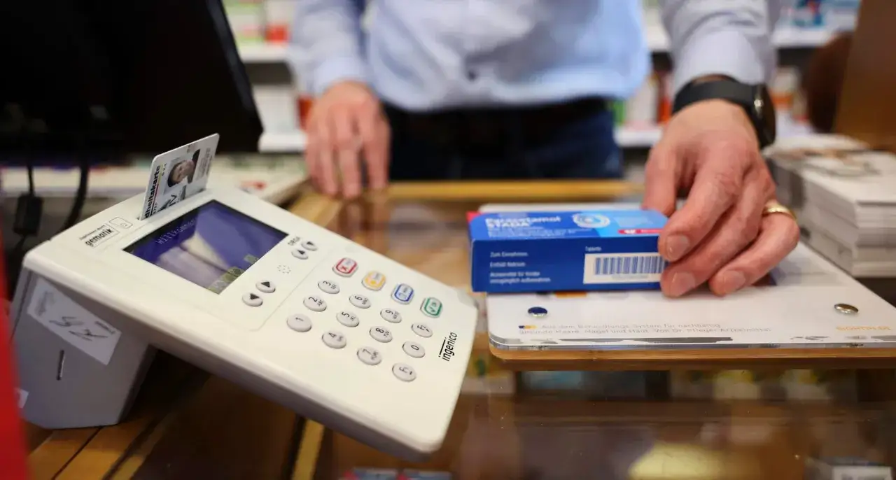 Krankenkassenkarte steckt in Terminal, Hand greift nach Medikamentenpackung.