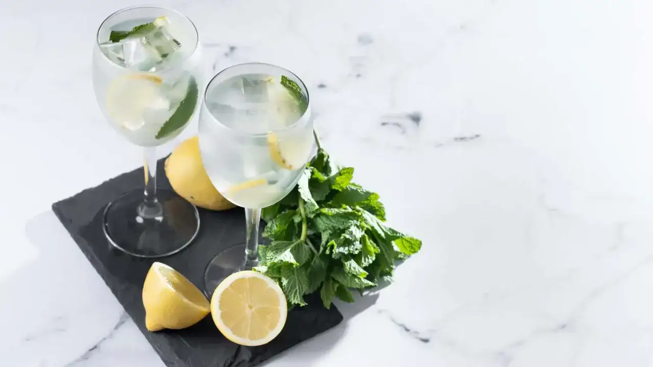 Dwa orzeźwiające drinki z prosecco, lodem, cytryną i miętą, prezentowane na czarnej desce z dodatkiem świeżych cytryn i gałązki mięty.