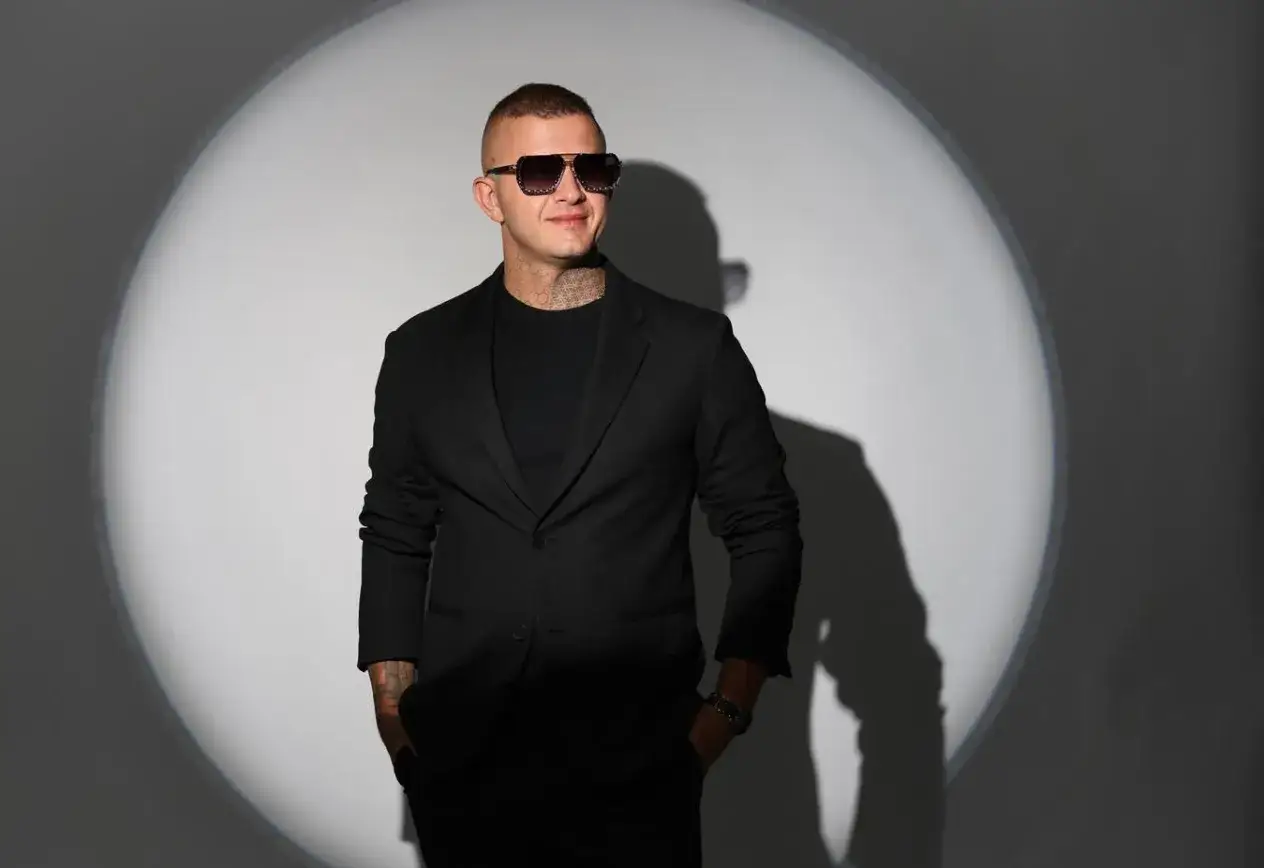 Mężczyzna w czarnym garniturze i okularach przeciwsłonecznych, z tatuażami na szyi. Jego uśmiech i postawa sugerują, że "ona działa na mnie jak" tekst piosenki.