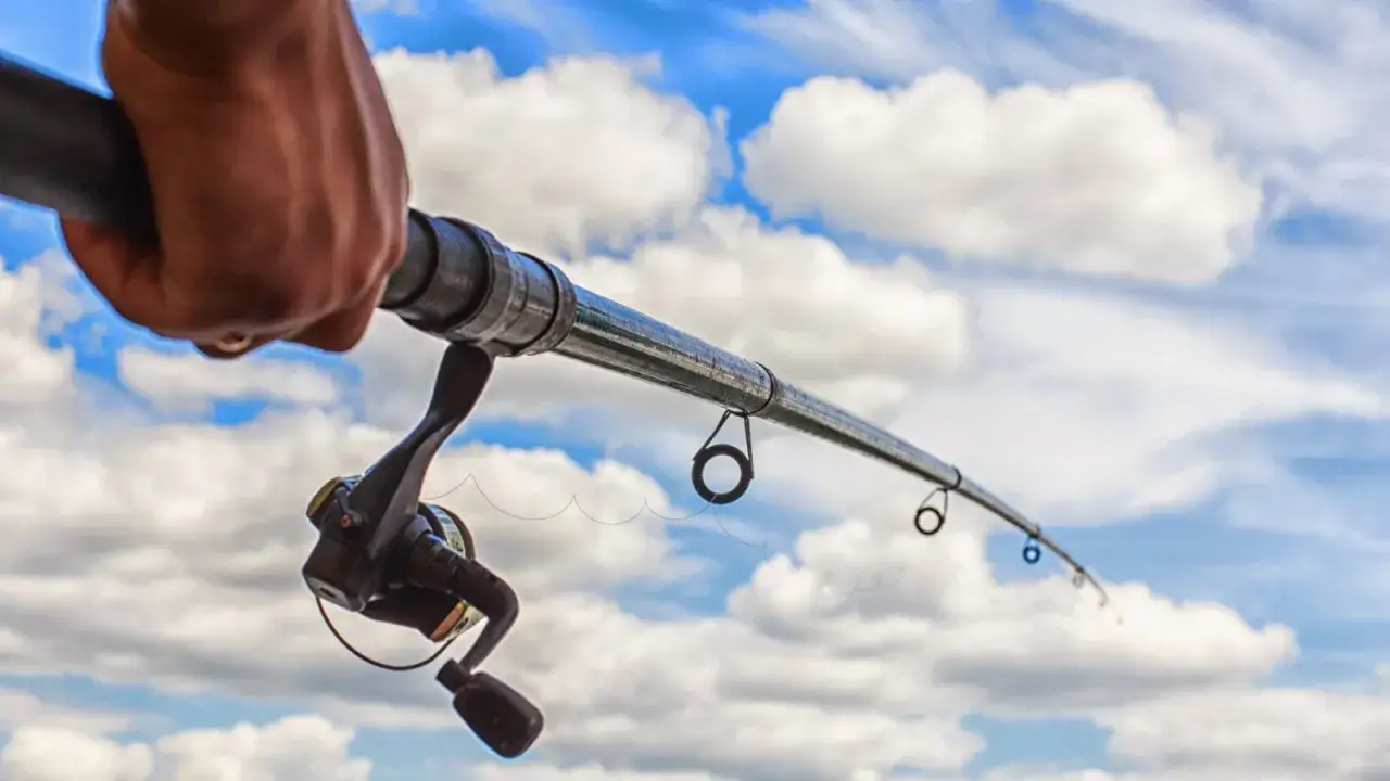 Dłoń trzyma wędkę z kołowrotkiem na tle błękitnego nieba z chmurami. Zastanawiasz się, ile kosztuje wędka?