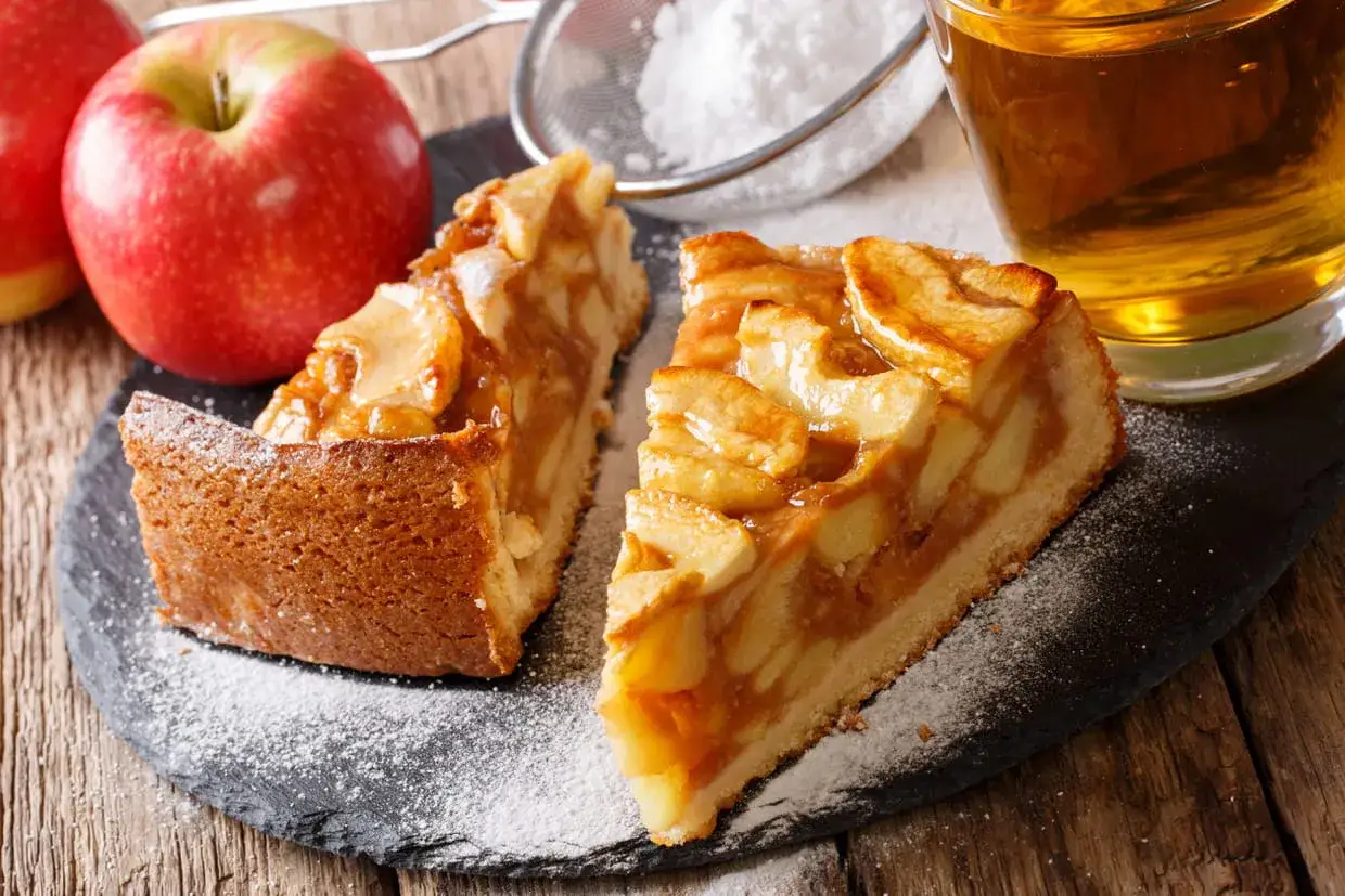 Deux parts de tarte aux pommes sur une ardoise, saupoudrées de sucre glace, avec des pommes et un verre de cidre.