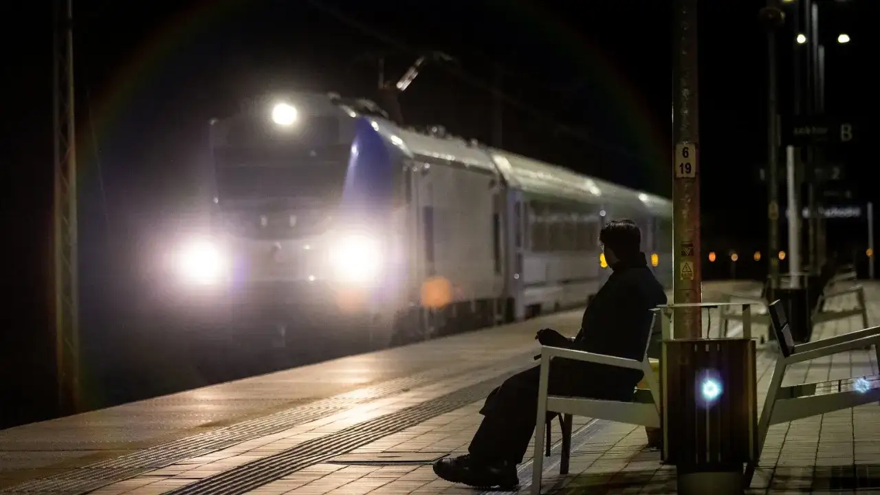 Osoba siedzi na ławce na peronie nocą, zbliża się pociąg z zapalonymi światłami.