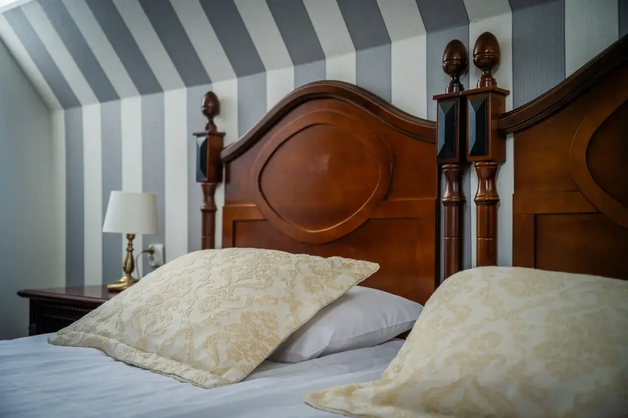 Eleganckie łóżko z drewnianym zagłówkiem i ozdobnymi poduszkami. Ciekawe, ile wynosi ryczałt za nocleg w takim miejscu?