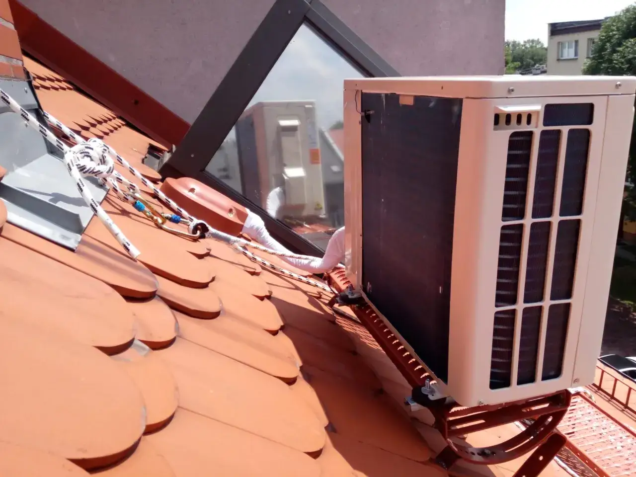 Klimatyzacja na dachu, zamontowana na czerwonej dachówce. Liny asekuracyjne zabezpieczają pracę instalatora.