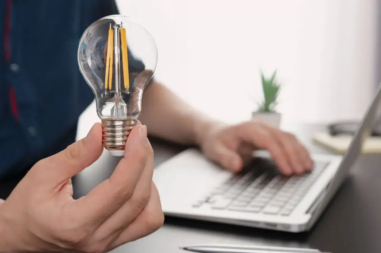 Dłoń trzyma żarówkę LED, symbolizującą pomysł na oszczędność energii, jak w taryfie c12a. W tle widać pracującego przy laptopie mężczyznę.