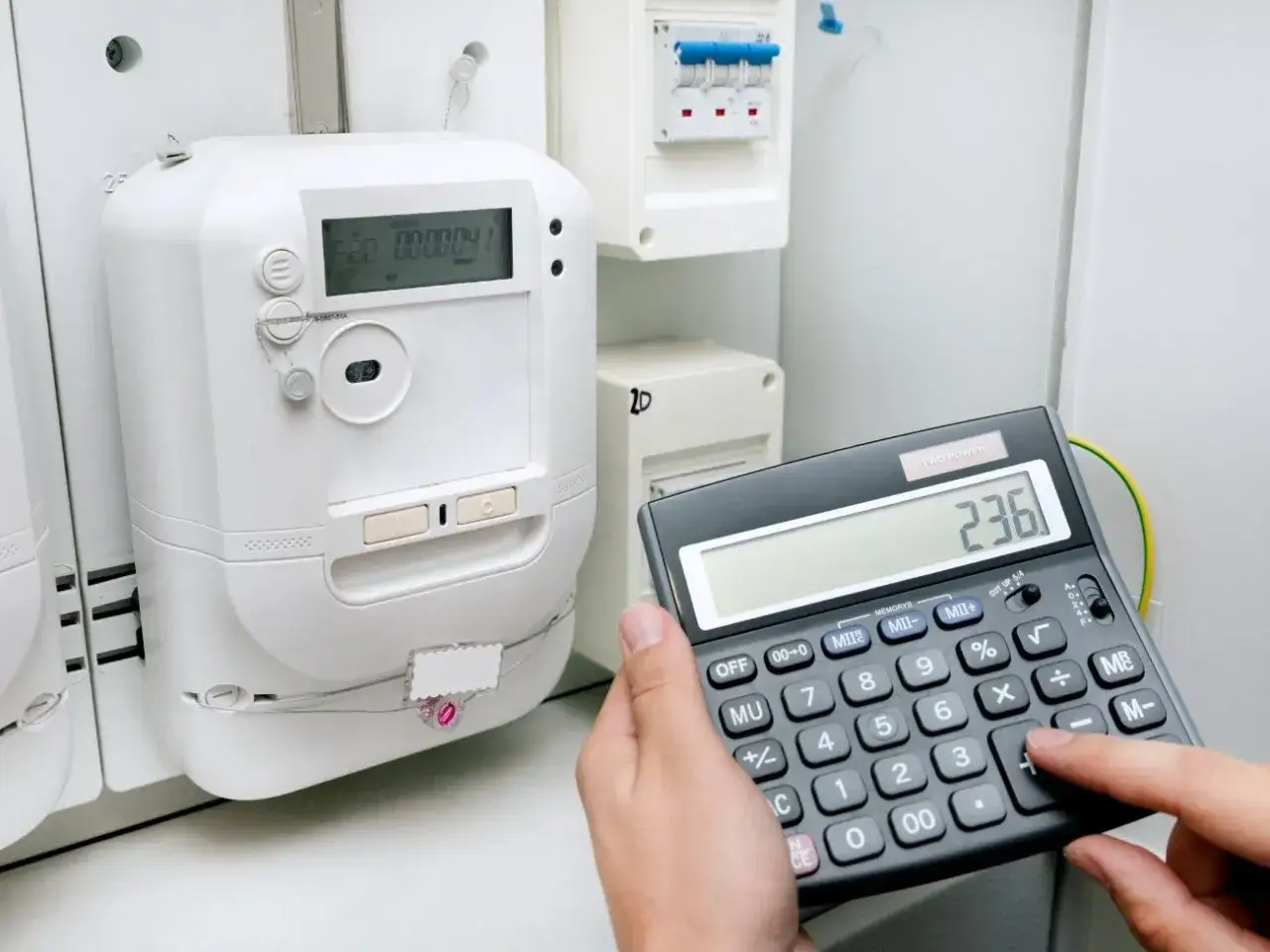 Dłoń z kalkulatorem przy liczniku energii elektrycznej.