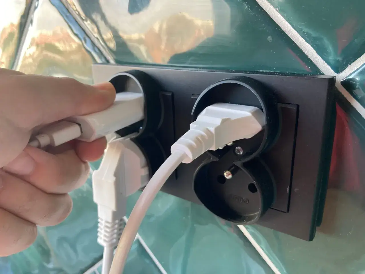 Dłoń podłącza biały kabel do gniazdka. W tle widać inne podłączone urządzenia, co może sugerować troskę o rachunki za prąd i potencjalną opłatę handlową PGE.