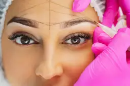 Nahaufnahme einer Frau, deren Augenbrauen mit Linien markiert werden, während eine Person in pinken Handschuhen mit einem Stift arbeitet.