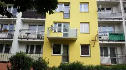 Żółty blok z dobudową balkonu na słupach. Widać okna, balustrady i anteny satelitarne.