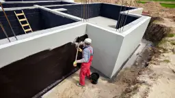 Budowlaniec maluje ciemną masą fundamenty domu.