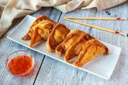 Crujientes wan tun frito dorados en una bandeja blanca, acompañados de salsa agridulce y palillos.