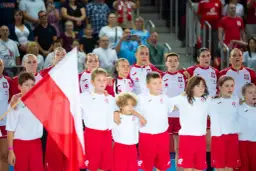 Młode zawodniczki i zawodnicy w strojach reprezentacji Polski, z biało-czerwoną flagą, przed meczem piłka ręczna kobiet mistrzostwa świata.