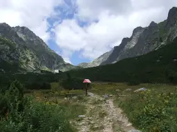 Kamienista ścieżka w górskiej dolinie z zielonymi zboczami i budynkiem w tle.