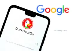 Na ekranie telefonu widoczny jest interfejs wyszukiwarki DuckDuckGo, która jest najlepszą wyszukiwarką internetową dbającą o prywatność. W tle rozmyty napis Google.