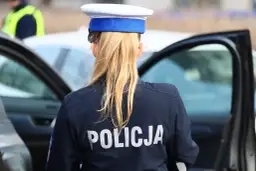 Policjantka w mundurze z napisem "POLICJA" na plecach, stojąca obok otwartych drzwi radiowozu. To jej dodatek funkcyjny.