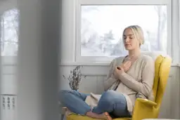 Kobieta z zamkniętymi oczami medytuje w żółtym fotelu, z rękami na piersi.
