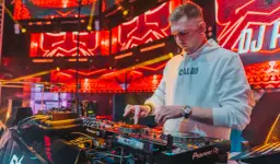 DJ w białej bluzie z kapturem gra na konsoli DJ-skiej na tle czerwonych świateł scenicznych.