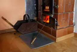 Piec kaflowy z otwartym paleniskiem, żarem i pogrzebaczem. Obok stoi metalowy kosz na węgiel. Pytanie: czy ekogroszkiem można palić w piecu kaflowym?