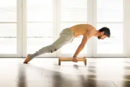 Hombre haciendo flexiones en paralelas de madera en un suelo claro, con luz entrando por ventanas al fondo.