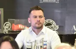 Grzegorz Collins, młody mężczyzna w białej koszuli, z tatuażem na szyi, siedzi przy stole. W tle widoczny pomarańczowy but sportowy.