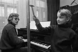 Zbigniew Wodecki gra na pianinie, a Paweł Wodecki trzyma młotek, jakby chciał uderzyć w klawisze.