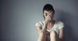 Kobieta w białej koszulce siedzi przy szarej ścianie, zakrywając twarz dłońmi. Czasem każdy potrzebuje chwili miansec, by zebrać myśli.