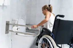 Kobieta w wózku inwalidzkim korzysta z uchwytów przy toalecie w łazience.