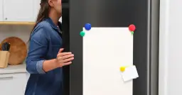 Kobieta w dżinsowej koszuli otwiera drzwi lodówki z pustą kartką i kolorowymi magnesami.