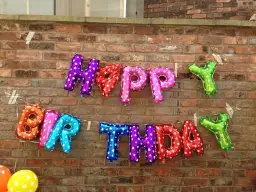 Luftballons in Buchstabenform bilden "HAPPY BIRTHDAY" an einer Ziegelwand.