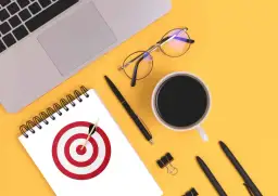 Na biurku leżą laptop, okulary, kubek kawy i notatnik z tarczą strzelniczą. To inspiracja, jak stworzyć personę.