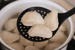 Gotowane pierogi na czarnej łyżce cedzakowej w garnku z wodą.