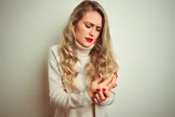 Kobieta z blond włosami trzymająca się za bolącą rękę, ubrana w beżowy golf.