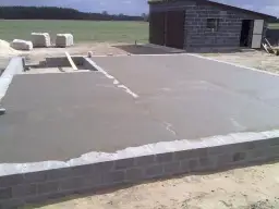Świeżo wylana płyta betonowa, z widoczną minimalną grubością chudziaka, na fundamencie z bloczków. W tle budynek gospodarczy.