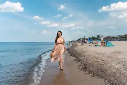 Kobieta w kwiecistej sukience spaceruje brzegiem morza. W tle plaża pełna ludzi, parawanów i domków – typowe sarbinowo atrakcje.
