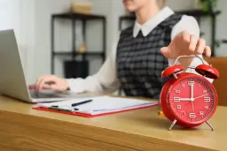 Kobieta wyłącza budzik, pracując przy laptopie. Czyżby to początek dnia w modelu 4 dniowy tydzień pracy?