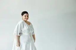 Kobieta w białej sukience z ozdobnym pasem, uśmiecha się do aparatu.