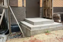 Solidny fundament pod schody z betonu, gotowy na dalsze prace budowlane. Obok widać materiały budowlane i fragment domu.