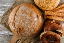 Różne rodzaje pieczywa: bochenek, paluszki sezamowe, obwarzanki i chleb z ziarnami.