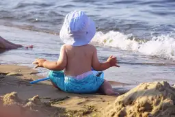 Niemowlak w kapelusiku i kąpielówkach z rekinem siedzi na piasku, patrząc na fale. Idealne miejsce, gdzie nad morze z niemowlakiem.