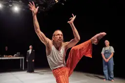Tancerz w pomarańczowych spodniach wykonuje dynamiczny ruch, prezentując sztukę na wynos. W tle widzimy innych performerów.