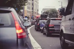 Korek w mieście, gdzie jest ruch lewostronny. Samochody stoją w rzędzie, widać znaki drogowe i budynki.